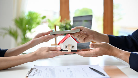 "Hands of a male client handing over money to a female real estate agent who is holding a small model house, symbolizing a house loan transaction. Documents and a pen are visible on the table in the foreground."